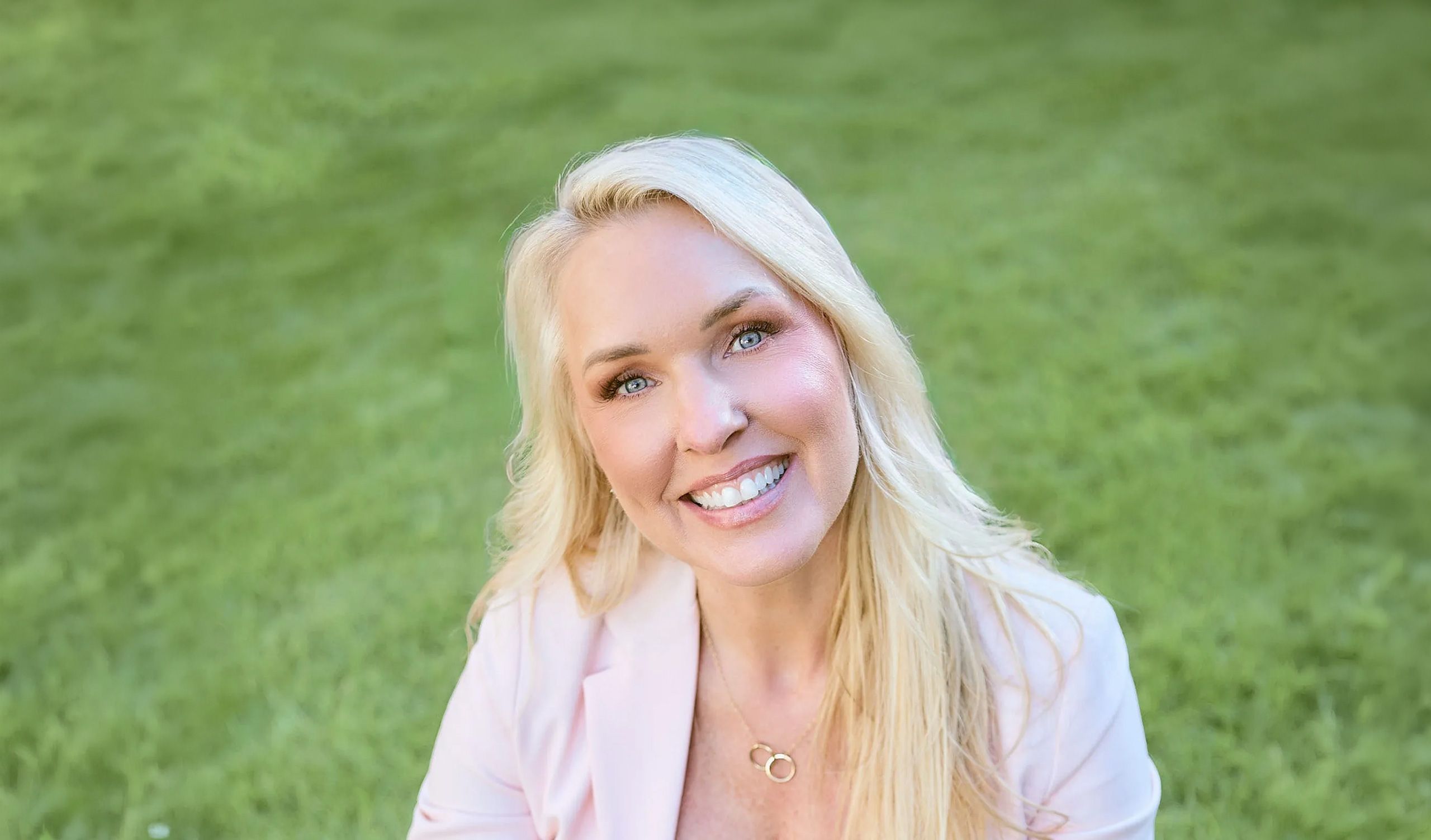 Dr. Karen Stewart in a pink blazer and jeans is sitting with a smile on her face against a green background.
