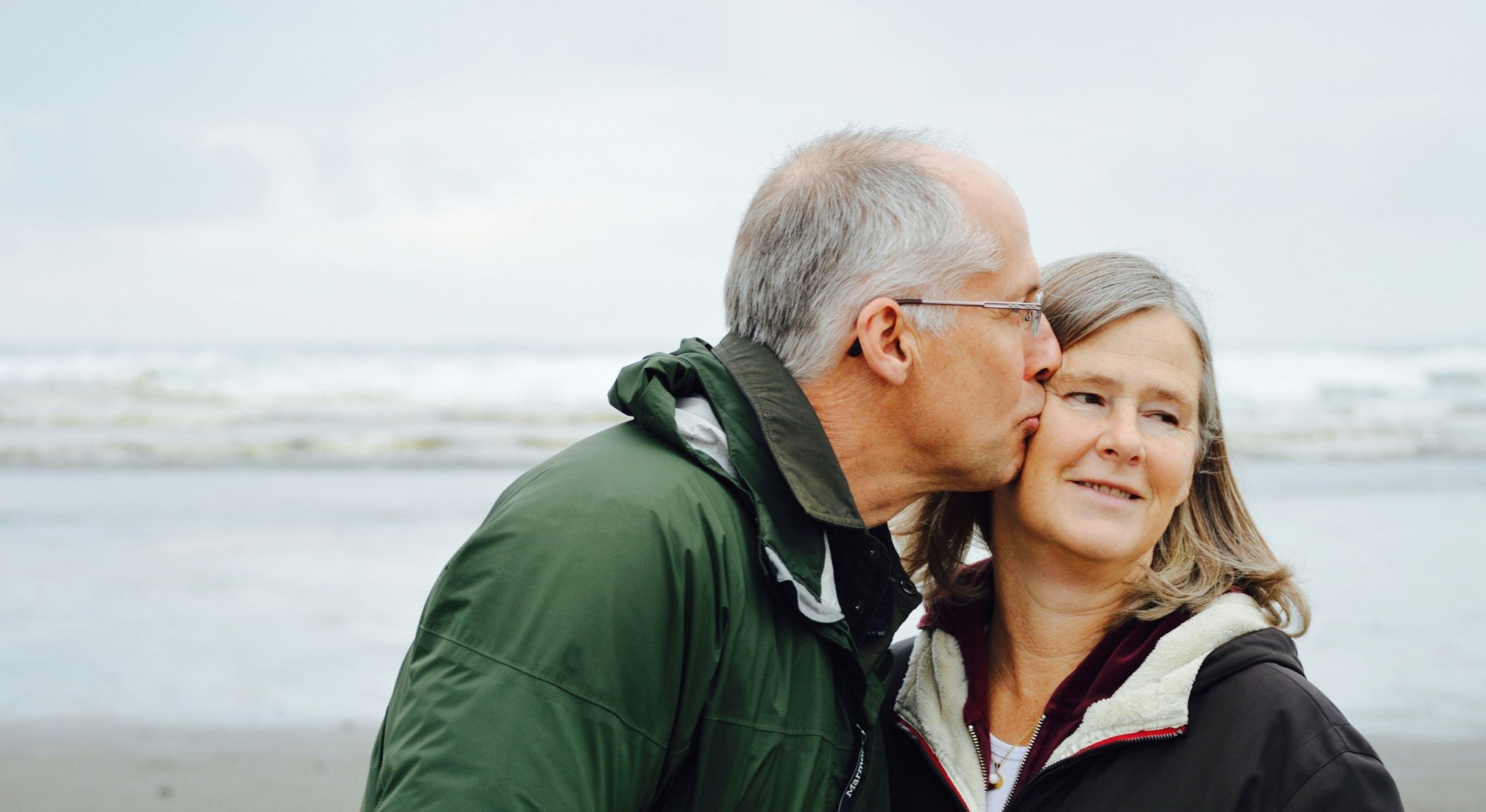 An older man affectionately kisses a woman on the cheek on a sandy beach by the ocean